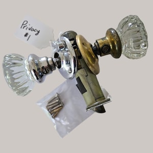 May include: A door knob set featuring two clear, fluted glass knobs with one chrome and one brass-toned metal base. The mechanism includes a latch assembly and a small plastic bag containing several metal screws. A white tag is attached with the text Privacy $1.