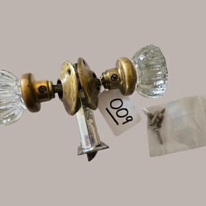 May include: A door knob set featuring two clear, fluted glass knobs with brass-toned metal bases and a central latch mechanism. A white tag with the handwritten number 009 is attached, and a small plastic bag contains three metal screws.