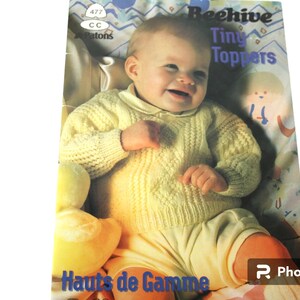 May include: A yellow knitted baby sweater with a cable knit pattern. The sweater is being worn by a baby sitting in a chair. The image is from a knitting pattern book titled "Beehive Tiny Toppers".