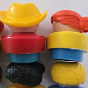 May include: Close-up of several colourful toy figures stacked together. The figures have various hair colours, including red, black, and yellow, and wear tops in red, yellow, and blue. One figure wears a yellow cowboy hat.