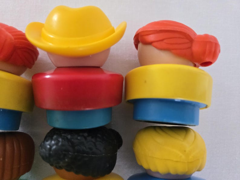May include: Close-up of several colourful toy figures stacked together. The figures have various hair colours, including red, black, and yellow, and wear tops in red, yellow, and blue. One figure wears a yellow cowboy hat.