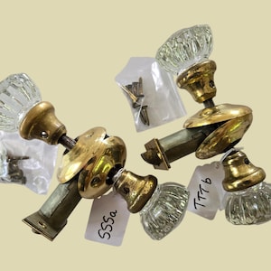 May include: Two sets of brass door knobs with clear, fluted glass handles. Each set includes a metal latch mechanism, a brass mounting plate, and a small plastic bag containing metal screws. One set is labeled 555a and the other is labeled TTTb.