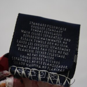May include: A dark blue rectangular fabric care label attached to a red and white patterned textile. The label features white printed text in English, French, and Spanish, including care instructions and the words STANDARD PILLOWCASE.