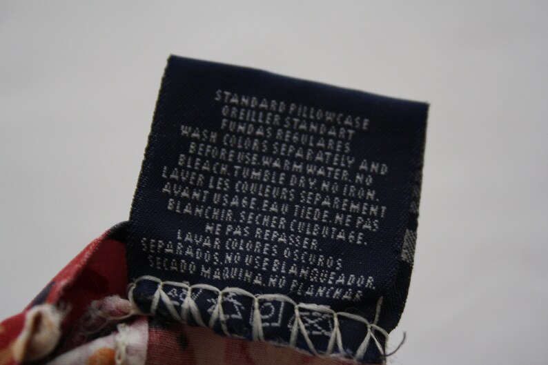 May include: A dark blue rectangular fabric care label attached to a red and white patterned textile. The label features white printed text in English, French, and Spanish, including care instructions and the words STANDARD PILLOWCASE.