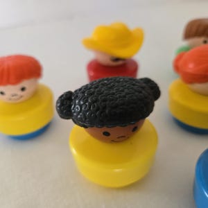 May include: A selection of vintage Fisher-Price Little People figures. The figures have yellow, blue, and red bases, with various hair colours and styles, including red, brown, and black. Some figures have hats.