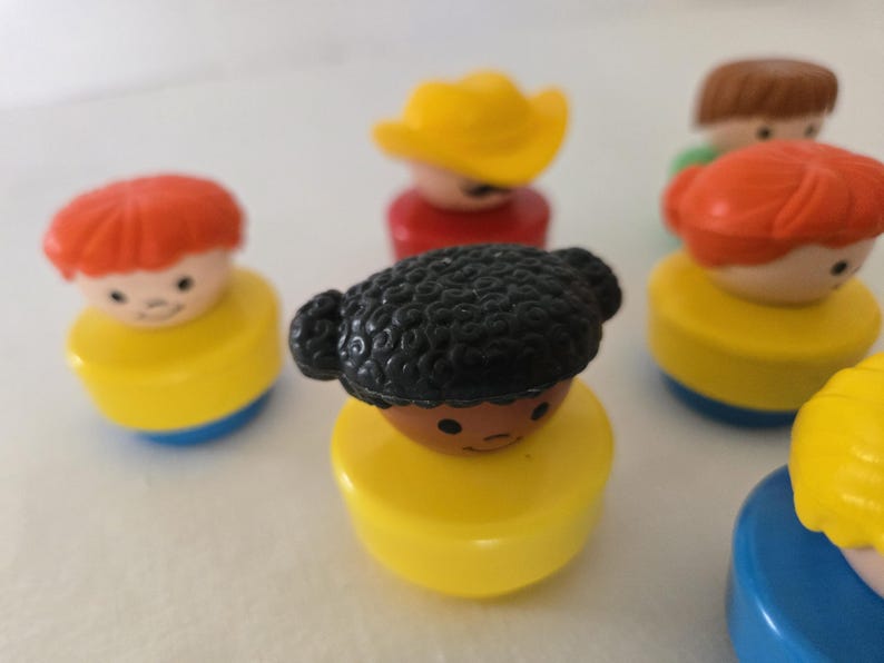 May include: A selection of vintage Fisher-Price Little People figures. The figures have yellow, blue, and red bases, with various hair colours and styles, including red, brown, and black. Some figures have hats.