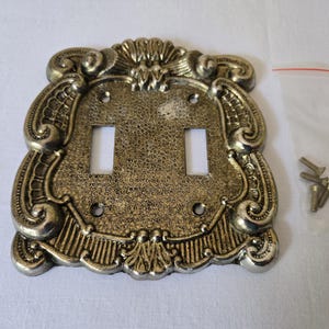 May include: A double light switch wall plate with an ornate, textured gold and silver finish. The rectangular plate features two vertical cutouts for switches and four screw holes. Four small metal screws are included in a clear plastic bag.