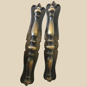 May include: A pair of antique bronze door handles. Each handle features an ornate, elongated backplate with a curved top. The handles are cylindrical with a rope-like detail and a rounded top. The finish has a warm, aged appearance.