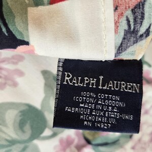 May include: A dark blue fabric clothing label with white text reading Ralph Lauren, 100% Cotton, Made in U.S.A., and RN 14927, attached to a floral patterned textile featuring pink, green, and black botanical illustrations on a cream background.