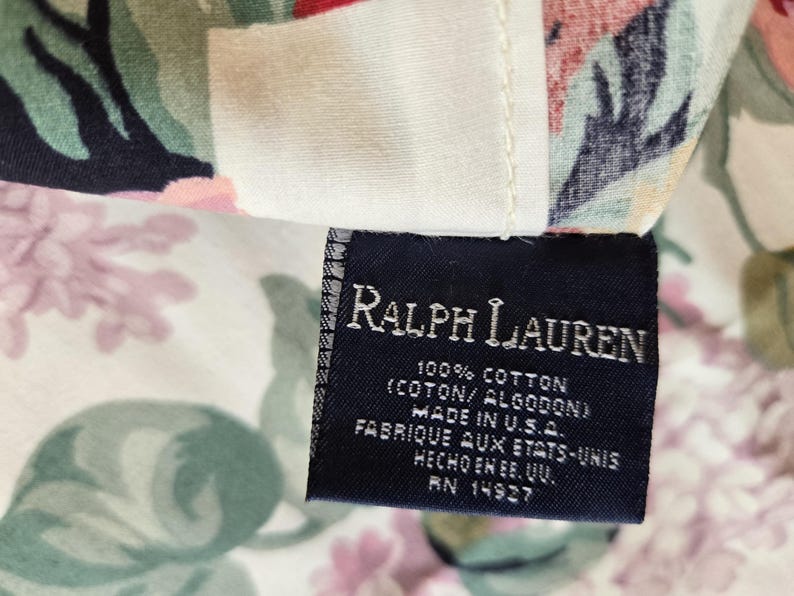 May include: A dark blue fabric clothing label with white text reading Ralph Lauren, 100% Cotton, Made in U.S.A., and RN 14927, attached to a floral patterned textile featuring pink, green, and black botanical illustrations on a cream background.