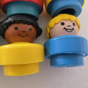 May include: Close-up of several vintage Fisher-Price Little People figures. The figures have smiling faces, with yellow, brown, and black hair. The figures have yellow, blue, and green bases. The figures are a classic toy.