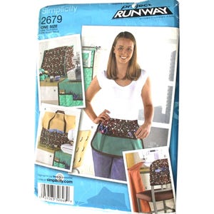 May include: Simplicity 2679 Project Runway sewing pattern envelope for a one-size utility apron with pockets. The cover features images of the apron in various fabric prints, including a brown pattern with safety pins and a solid green fabric.
