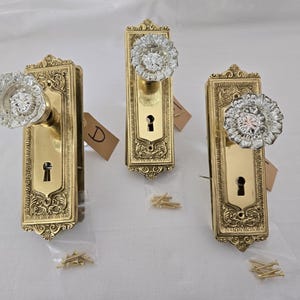 Peut inclure: Trois boutons de porte en laiton antique avec des détails ornés et des boutons en cristal de verre transparent. Chaque bouton est monté sur une plaque rectangulaire avec un trou de serrure.