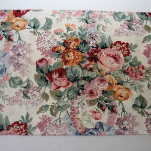 May include: A rectangular fabric placemat featuring a cream-coloured background with a dense, colourful floral pattern. The design includes clusters of pink, orange, and red roses, purple lilac sprigs, and green foliage, accented by light blue ribbon motifs.