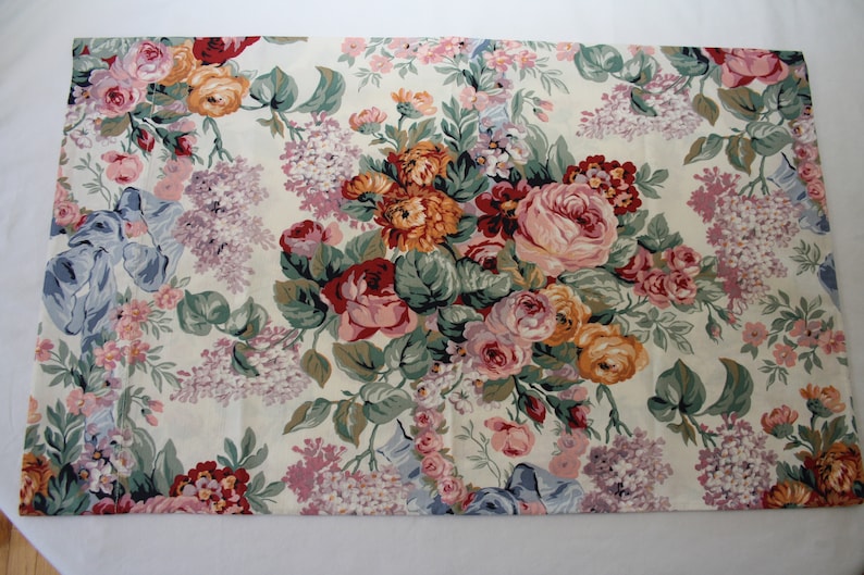 May include: A rectangular fabric placemat featuring a cream-coloured background with a dense, colourful floral pattern. The design includes clusters of pink, orange, and red roses, purple lilac sprigs, and green foliage, accented by light blue ribbon motifs.
