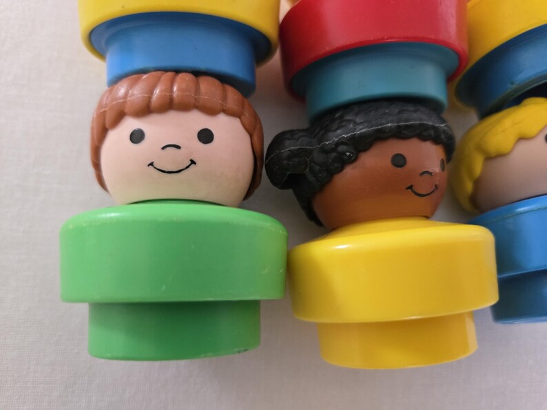 May include: Close-up of several colourful, vintage toy figures. Each figure has a smiling face and a different hair colour, including brown, black, and blonde. The figures are stacked on top of each other, with a variety of bright colours.