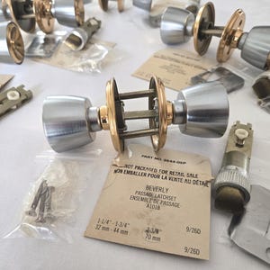 May include: A collection of silver and gold-coloured door knobs and associated hardware. The image shows complete knobs, latches, and screws. The packaging includes the text "BEVERLY PASSAGE LATCHSET" and measurements of 32 mm - 44 mm.