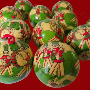 May include: A collection of round, glossy Christmas baubles featuring a repeating illustration of toy soldiers in red and green uniforms playing musical instruments like drums and horns on a cream-coloured background with green grass accents.