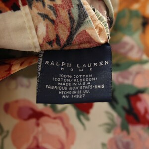 May include: A Ralph Lauren Home fabric tag attached to a floral patterned textile. The dark blue rectangular label features white text reading Ralph Lauren Home, 100% Cotton, Made in U.S.A., and RN 14927, set against a cream background with red and green flowers.