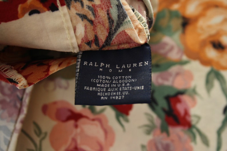 May include: A Ralph Lauren Home fabric tag attached to a floral patterned textile. The dark blue rectangular label features white text reading Ralph Lauren Home, 100% Cotton, Made in U.S.A., and RN 14927, set against a cream background with red and green flowers.