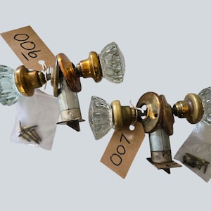 May include: Two sets of brass door knobs with clear, faceted glass handles. Each set includes a metal latch mechanism and a small plastic bag containing mounting screws. One tag is labeled 900 and the other is labeled 007 in black ink.