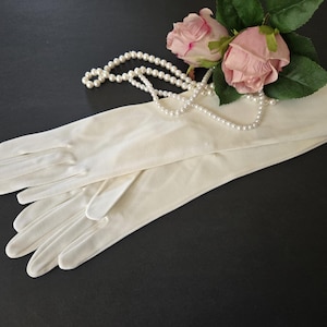 May include: A pair of long, off-white opera gloves made of a smooth, opaque fabric. The gloves are positioned alongside a strand of white faux pearl beads and two artificial pink roses with green leaves, creating a coordinated set of accessories.