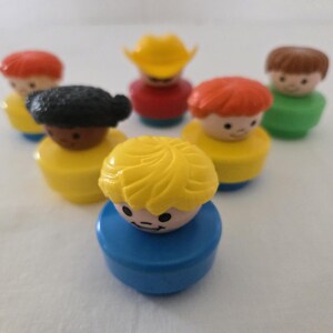 May include: A selection of vintage Fisher-Price Little People figures. The figures have different hair colours, including red, brown, and yellow, and wear outfits in yellow, red, and green. One figure wears a yellow cowboy hat. The toys are on a white surface.