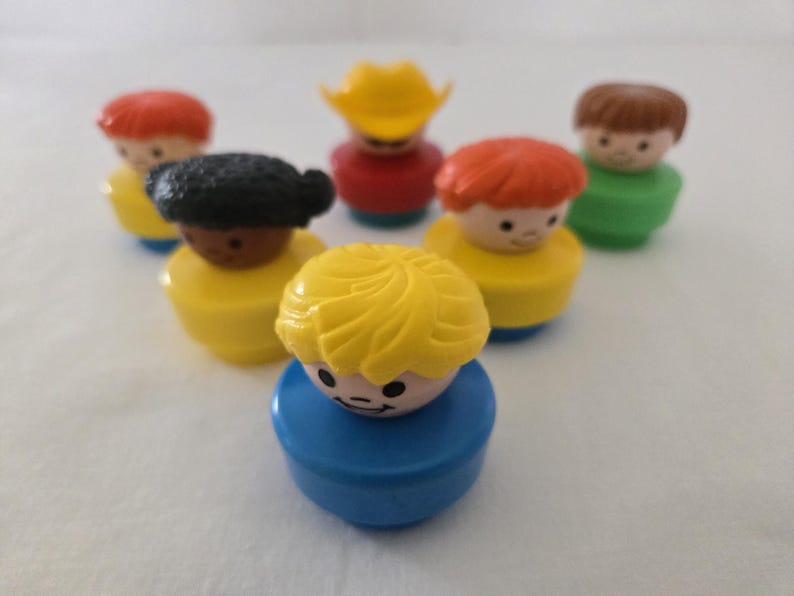May include: A selection of vintage Fisher-Price Little People figures. The figures have different hair colours, including red, brown, and yellow, and wear outfits in yellow, red, and green. One figure wears a yellow cowboy hat. The toys are on a white surface.