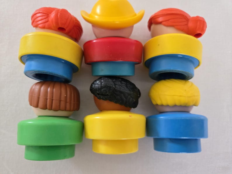 May include: A selection of vintage Fisher-Price Little People figures. The figures have different hair colours, including red, brown, black, and yellow. The figures have yellow, red, blue, and green clothing. The figures are stacked together.