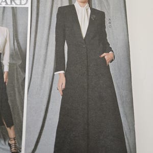 May include: A long, dark gray coat with a single-button closure and a notched collar. The coat is paired with a white blouse and a brooch. The model is wearing black heels. The coat has a tailored fit and a classic design.