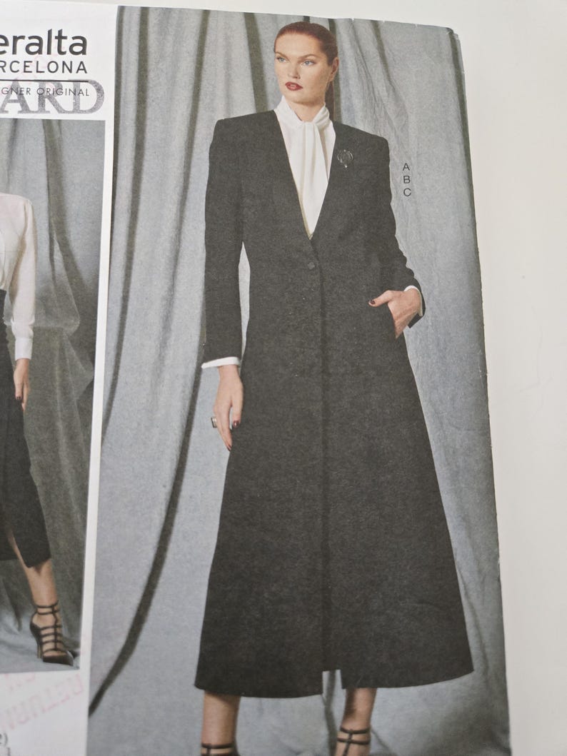 May include: A long, dark gray coat with a single-button closure and a notched collar. The coat is paired with a white blouse and a brooch. The model is wearing black heels. The coat has a tailored fit and a classic design.
