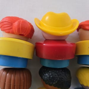 May include: A close-up of several colourful toy figures stacked on top of each other. The figures have different coloured hats, hair, and body sections. The colours include yellow, red, blue, brown, and black. The figures are designed to be stacked.