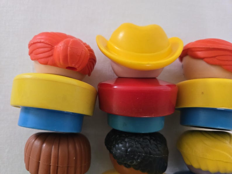 May include: A close-up of several colourful toy figures stacked on top of each other. The figures have different coloured hats, hair, and body sections. The colours include yellow, red, blue, brown, and black. The figures are designed to be stacked.