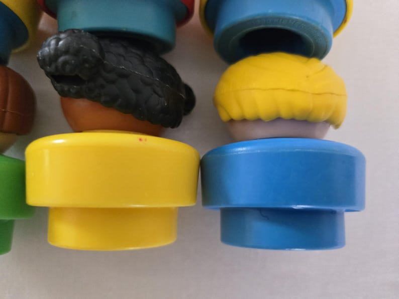 May include: Close-up of colourful plastic toy figures. The figures have yellow, blue, and brown bases with different coloured heads, including yellow and black hair. The toys are likely designed for children.