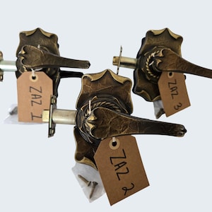 May include: Three antique bronze door handles with ornate detailing. Each handle has a lever and a decorative backplate. Attached to each handle is a brown tag with the text "ZAZ" and a number. The handles are displayed against a light background.