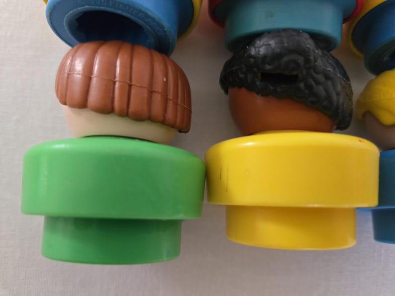 May include: Close-up of several vintage toy figures. The figures have brown, black, and yellow hair, with green and yellow bases. The figures are stacked on top of each other, with blue and red tops.