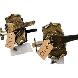 May include: Two antique brass door handles with ornate leaf detailing. Each handle has a dark brown lever and is attached to a metal plate. Tags labelled "CH 7" and "CH 8" are attached.