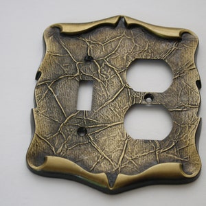 May include: Decorative light switch and outlet cover with a textured, aged bronze finish. The cover has a switch opening and two outlet openings. The edges are trimmed with a gold-coloured accent, adding elegance to the design.