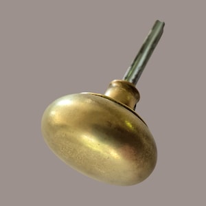 May include: A brass door knob with a smooth, rounded, oval-shaped handle and a metallic finish. The knob features a central cylindrical neck that connects to a long, slender, square-profile metal spindle extending outward from the back of the piece.