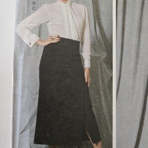 May include: A woman's outfit featuring a white blouse with a high collar and long sleeves, paired with a dark gray skirt. The skirt has a front slit. The outfit is completed with black strappy heels.