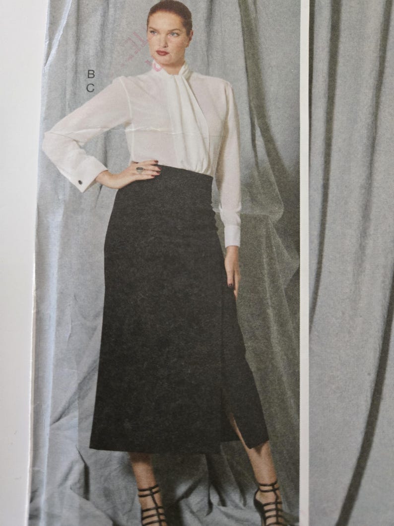 May include: A woman's outfit featuring a white blouse with a high collar and long sleeves, paired with a dark gray skirt. The skirt has a front slit. The outfit is completed with black strappy heels.
