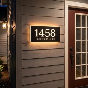 Peut inclure: Une plaque d'adresse rectangulaire noire fixée sur un bardage gris horizontal à côté d'une porte rouge foncé. La plaque affiche les chiffres blancs 1458 et le texte CALIFORNIA DR, rétroéclairés par une lueur chaude contre le mur.