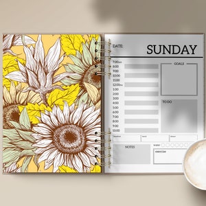 Puede incluir: Agenda abierta con un dise&ntilde;o de girasoles en la p&aacute;gina izquierda y un horario del domingo en la derecha. Incluye un bol&iacute;grafo con el texto "happiness looks gorgeous on you" y una taza de caf&eacute;.