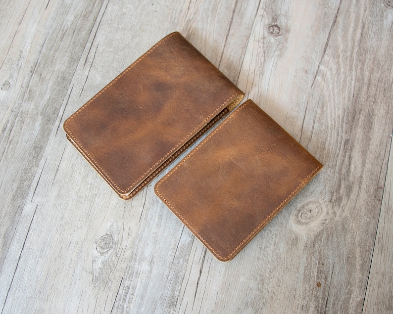 Pu&ograve; includere: Due portafogli in pelle marrone con bordi cuciti. I portafogli sono disposti su una superficie in legno chiaro.
