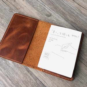 May include: Open brown leather notebook cover with a pocket on the left side. A white notebook with Japanese text and a hand-drawn illustration is inside. The cover is open on a wooden surface.