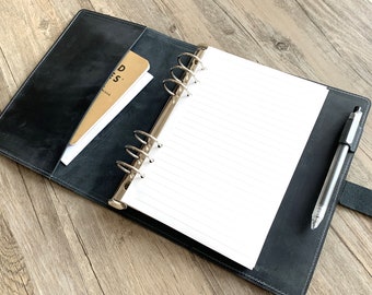 May include: A black leather bound planner with a silver ring binder. The planner is open to a blank page with lined paper. A pen and a small notebook are in the pockets of the planner.