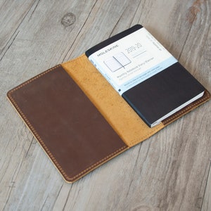 Puede incluir: Una cubierta de cuaderno de cuero marrón con un interior de cuero amarillo. Un cuaderno Moleskine negro con el texto "2019-20 Monthly Notebook Diary Planner" está dentro de la cubierta.