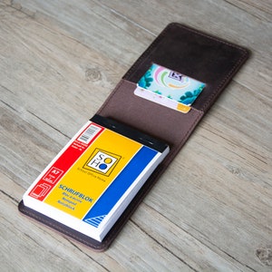 Pu&ograve; includere: Una copertina per quaderno in pelle marrone con un piccolo blocco note all'interno. Il blocco note ha una copertina gialla e blu con il testo "SCHRIFFBLOK" e "School Office Home" stampato su di essa.