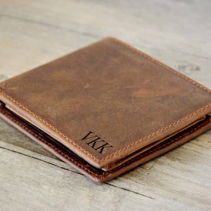 Puede incluir: Una cartera de cuero marrón con las iniciales "VKK" grabadas en la parte delantera. La cartera es rectangular y parece estar hecha de cuero envejecido, con costuras visibles alrededor de los bordes. La cartera está cerrada.