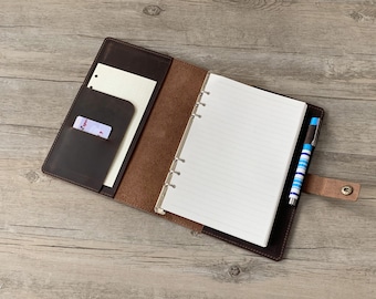 May include: A brown leather bound journal with a pen and a card holder. The journal has a ring binder and lined pages.
